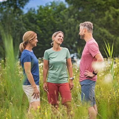 Drei Personen in sommerlicher Freizeitkleidung stehen in einer Blumenwiese mit hohem Gras und Blumen, umgeben von grünen Bäumen unter blauem Himmel.