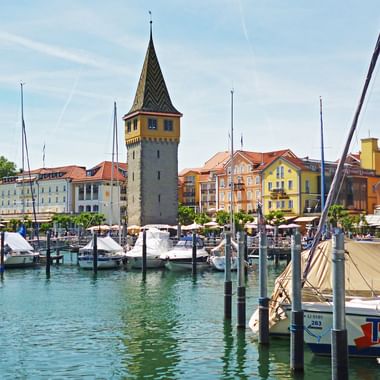 Lindau harbor at Lake Constance with the historic Mangturm tower, colorful buildings, sailboats, and turquoise water under blue sky.