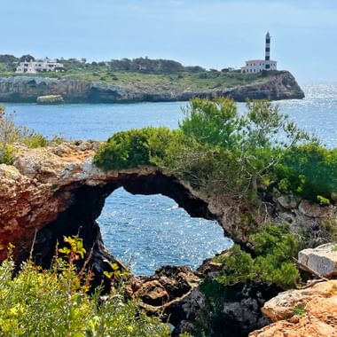 Natürlicher Felsbogen umrahmt von grüner Vegetation mit Blick auf türkisfarbene Bucht. Leuchtturm auf Felsenhalbinsel und weiße Gebäude im Hintergrund.
