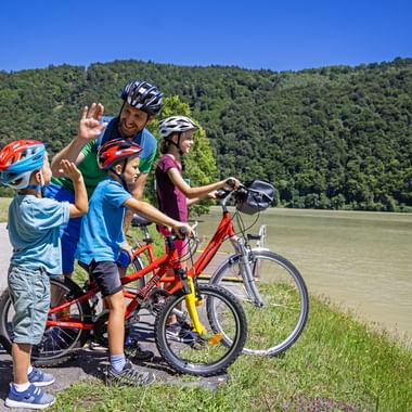 Family of four with bicycles giving high-fives by the Danube River. Two children and two adults wearing helmets with forested hills in background.