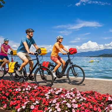 Three cyclists with panniers riding along a lakeside promenade with colorful flowers. Lake Garda and mountains visible in the background.