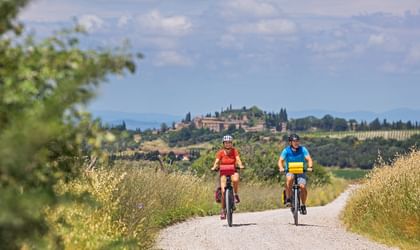 Zwei Radfahrer auf einem Schotterweg durch goldene Felder im Val d'Orcia, Toskana. Ein Bergdorf mit Zypressen ist in der Ferne sichtbar.