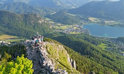 Eine Gruppe Wanderer steht rund um ein Gipfelkreuz auf einem Berg, Panoramablick auf See, Berge, sonniges Wetter