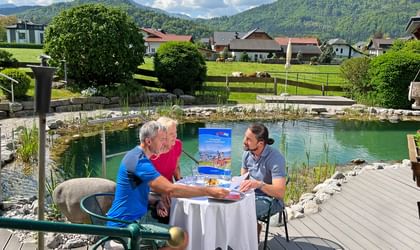 Three people at white table by natural swimming pond discussing travel brochures. Alpine village and forested mountains in background.
