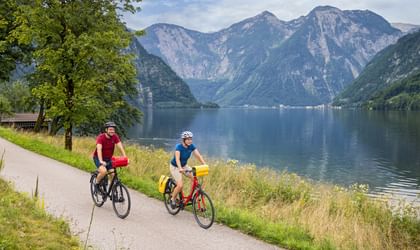 Two cyclists riding on a paved path along Lake Hallstatt with mountains in the background and calm blue water beside the trail.