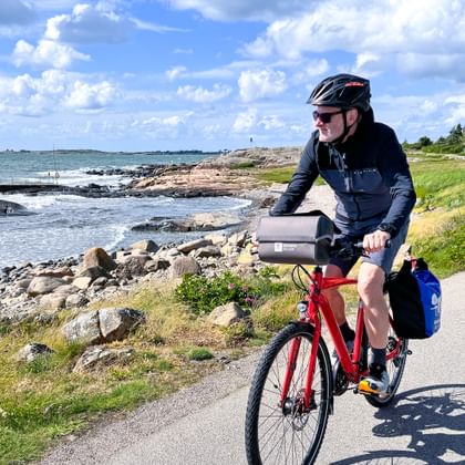 Cyclist with helmet riding red bike on coastal path along Swedish west coast near Varberg, with rocky shoreline and blue sea under cloudy sky.