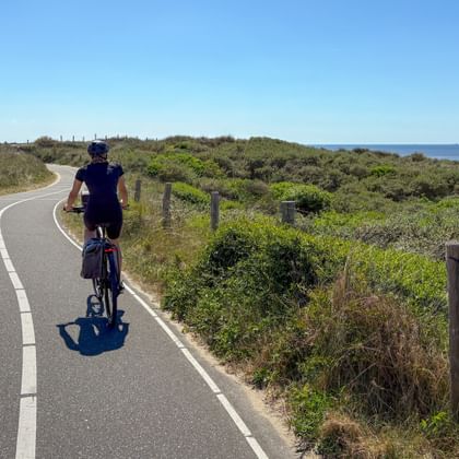 Cyclist riding on paved coastal bike path with green vegetation and ocean view. Clear blue sky above, wooden fence posts along the path.