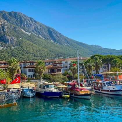Hafen von Akyaka mit bunten Booten unter türkischen Flaggen im türkisfarbenen Wasser. Weiße Häuser mit Palmen und grüner Berg unter blauem Himmel.