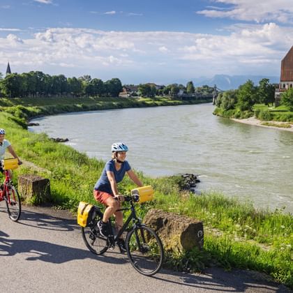 Three cyclists with yellow panniers riding along the Salzach River path near Laufen. The baroque Stiftskirche church is visible across the river.