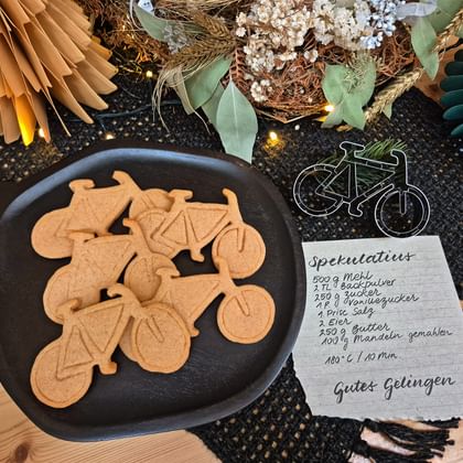 Black plate with bicycle-shaped cookies next to a handwritten recipe card and bike decoration. Festive wreath and paper fans in background.