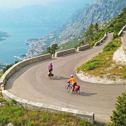 Two cyclists on a winding road with a view of the Bay of Kotor surrounded by mountains