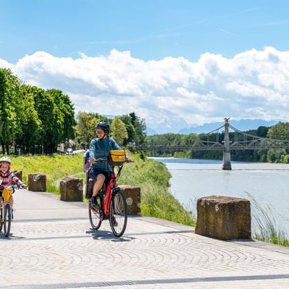 Adult and child cycling on paved riverside path near Oberndorf. Bridge visible across river, trees line the path under blue sky with clouds.