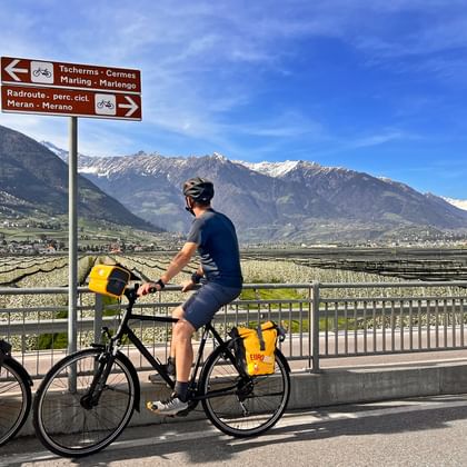 Radfahrer mit gelben Packtaschen am Etschradweg mit Blick auf schneebedeckte Berge. Wegweiser zeigen nach Tscherms, Marling und Meran.