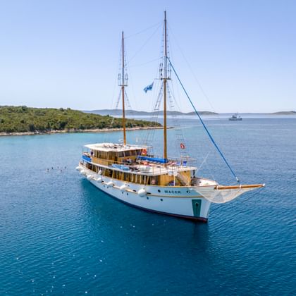 Weißes Motorsegelschiff MS Macek mit zwei Masten vor Anker in türkisfarbenem Adriatischem Meer nahe grüner kroatischer Küste.