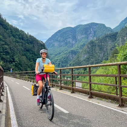 Radfahrerin mit gelben Packtaschen auf asphaltiertem Weg mit Metallgeländer, umgeben von bewaldeten Bergen im Alpental.