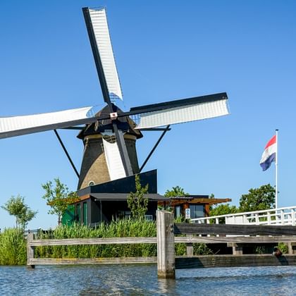 Traditional Dutch windmill with white sails at Kinderdijk beside a canal. A white wooden bridge with Dutch flag and people crosses the waterway.
