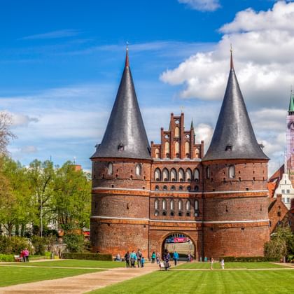 Historic Holstentor gate with twin towers in Lübeck, surrounded by green lawn and trees. Church spires and historic buildings visible in background.