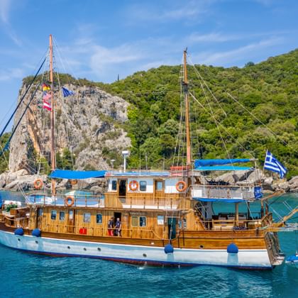 Traditionelles Holzsegelschiff MS Panagiota mit blauem Sonnensegel in türkisfarbener Bucht. Grüne bewaldete Hügel erheben sich aus dem Wasser.