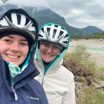 Two smiling cyclists in helmets and rain jackets taking a selfie by the Isar River with mountains and forest in the background.