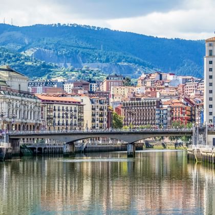 River view of Karlsbad with historic buildings, a bridge, and forested mountains in the background under a partly cloudy sky.