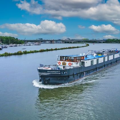 Flusskreuzfahrtschiff MS Magnifique II fährt auf breiter Wasserstraße unter blauem Himmel mit weißen Wolken. Marina und grüne Bäume am linken Ufer.