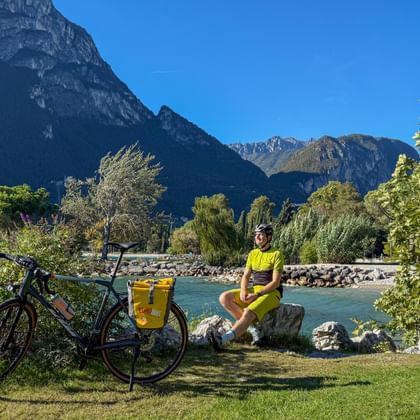Cyclist in yellow sitting on grass by turquoise lake with touring bike. Mountains and trees frame the scenic lakeside promenade in Riva.