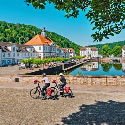 Zwei Radfahrer mit roten Taschen fahren am Barockhafen in Bad Karlshafen entlang, mit historischen weißen Gebäuden und einer Brücke im ruhigen Wasser.