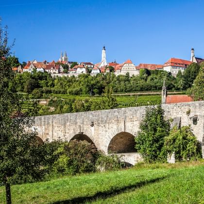 Historische Steinbrücke mit Bögen im Vordergrund, mittelalterliche Stadt Rothenburg ob der Tauber mit rotgedeckten Gebäuden und Kirchtürmen auf Hügel.