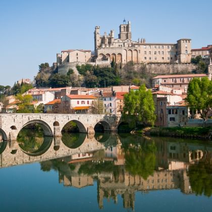 Steinbrücke mit mehreren Bögen über einem ruhigen Fluss in Béziers, mit der Kathedrale Saint-Nazaire auf dem Hügel und Spiegelungen im Wasser.