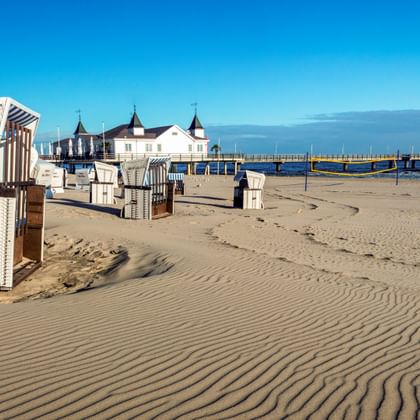 Sandstrand mit traditionellen Strandkörben und der Seebrücke Ahlbeck, die sich in die Ostsee auf Usedom erstreckt, unter klarem blauen Himmel.