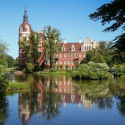 Rotes Backsteinschloss Muskau mit Uhrenturm spiegelt sich im See, umgeben von grünen Bäumen im Fürst-Pückler-Park unter blauem Himmel.