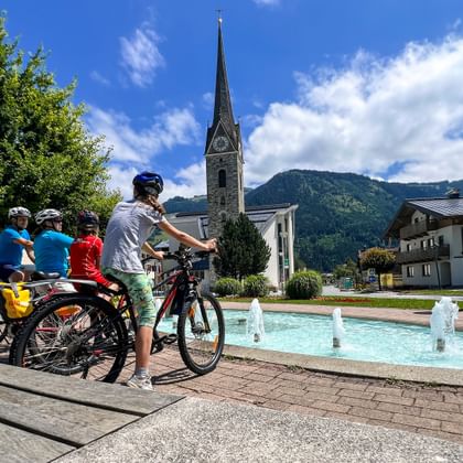 Family of four cycling past a fountain in Maishofen village square, with a church spire and mountains in the background under blue sky.