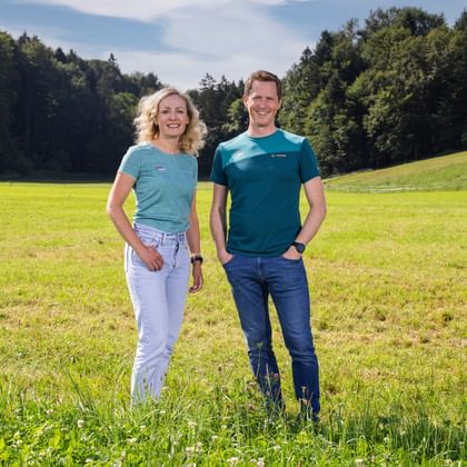 Two people Verena Sonnenberg und Thomas Schmid standing in a green meadow with forest in the background. Both wear blue outdoor shirts and smile at the camera.