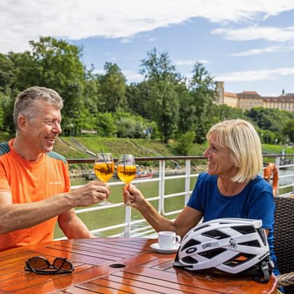 Couple toasting with drinks on ship terrace along the Danube. Cycling helmets on table, green riverbank and historic buildings in background.