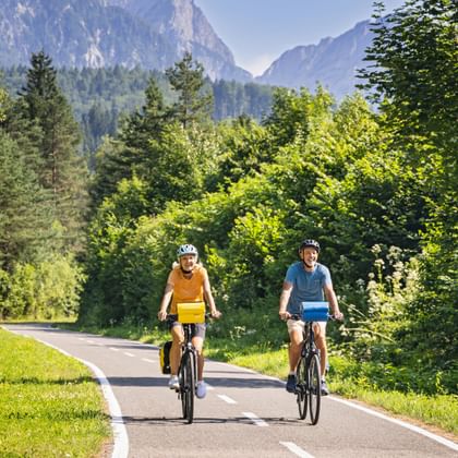 Two cyclists riding on paved path through Kanaltal valley near Tarvis, surrounded by green forest with mountain peaks in background.