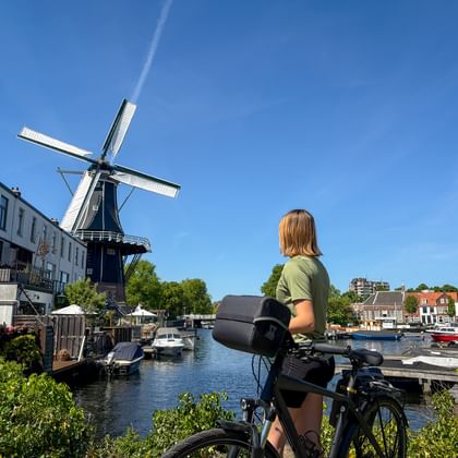 Radfahrerin mit Fahrrad betrachtet die historische Windmühle De Adriaan am Fluss Spaarne in Haarlem, mit Booten und traditionellen Gebäuden.