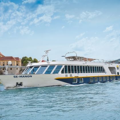 White river cruise ship MS Manon sailing on the Danube near Dürnstein with blue baroque church tower and hills in background.