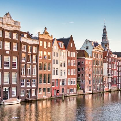 Reihe bunter historischer Grachten­häuser entlang Amsterdams Damrak mit Kirchturm im Hintergrund unter blauem Himmel.