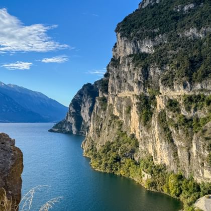 Panoramic view from Strada del Ponale showing steep cliffs, blue Lake Garda waters, and distant mountains under a clear sky.