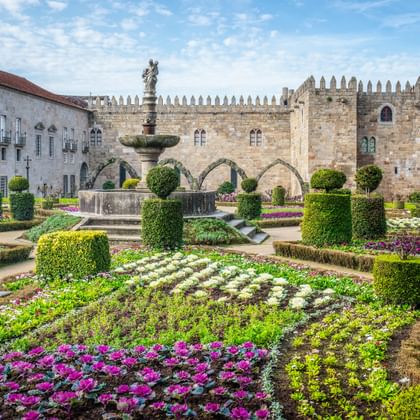 Jardim de Santa Barbara in Braga with colorful flower beds, sculpted hedges, and a fountain. Medieval stone buildings and walls in background.