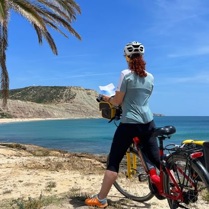 Radfahrerin mit Helm und Karte steht mit Tourenrad bei Palmen an einer Sandbucht mit türkisem Wasser und Hügeln.