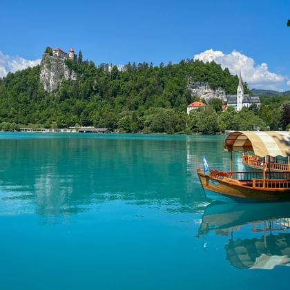 Wooden boat on turquoise Lake Bled with forested hill, Bled Castle, and white church tower. Mountains and blue sky with clouds in background.