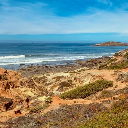 Zerklüftete Küstenlandschaft bei Porto Covo mit rotbraunen Klippen, grüner Vegetation und blauem Atlantik mit weißen Wellen und kleinen Inseln.