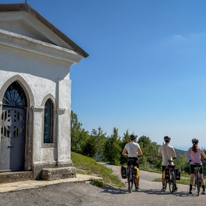 Three cyclists on a hilltop road beside a small white chapel with Gothic windows near Canebola, overlooking forested hills and distant sea.