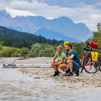 Two cyclists sitting on a pebble beach by the Isar River with touring bikes. Mountains and forest visible in the background under cloudy sky.