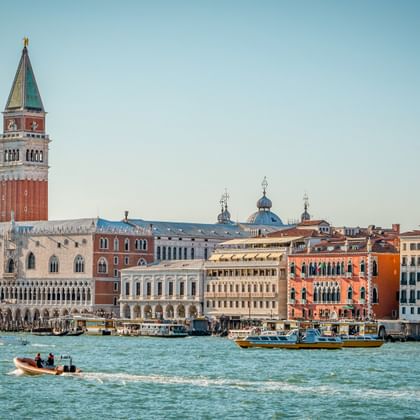 Venedigs Uferpromenade mit Markusturm und bunten historischen Gebäuden. Boote fahren durch die türkisfarbene Lagune unter blauem Himmel.