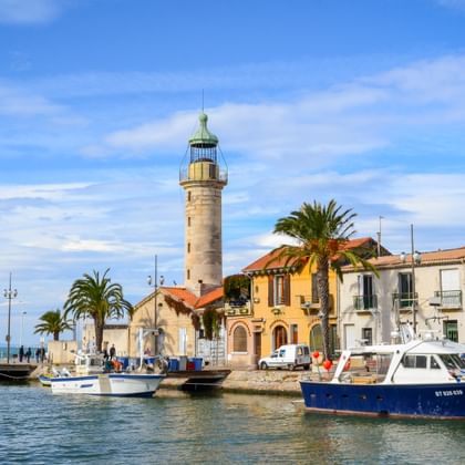 Stone lighthouse at Le Grau-du-Roi harbor with fishing boats, Mediterranean houses, palm trees and blue sky in Camargue, Provence.