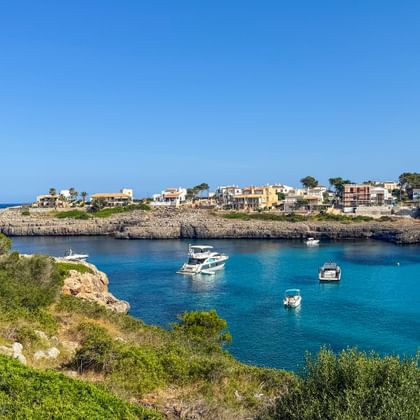 Turquoise bay with boats anchored in calm waters near Porto Colom, Mallorca. Rocky coastline with green vegetation and Mediterranean houses visible.