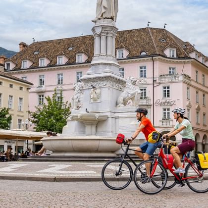 Zwei Radfahrer fahren am weißen Monumentbrunnen am Waltherplatz in Bozen vorbei, mit historischen pastellfarbenen Gebäuden und Straßencafés.