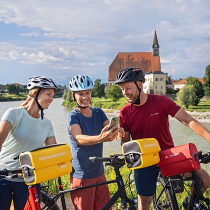 Three cyclists with helmets and handlebar bags viewing a smartphone by the Salzach River near Oberndorf, with a church in the background.